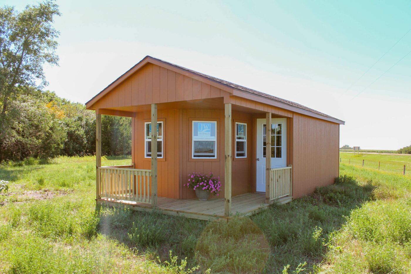 Orange cabin-style shed with a small covered porch, white door, and windows, set in a grassy rural area with trees in the background.