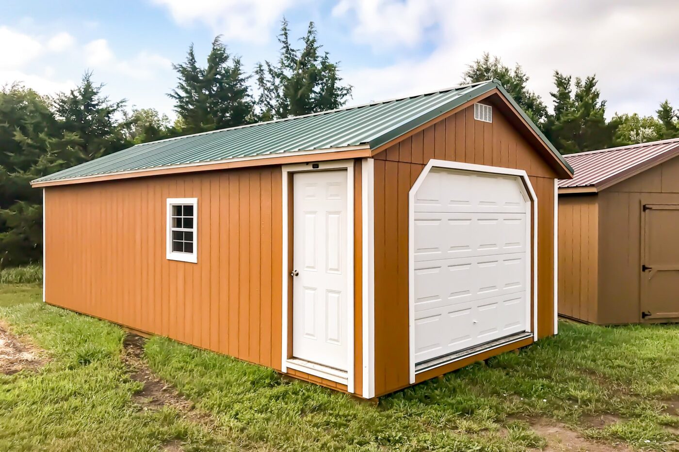 Wooden garage with an orange exterior, green metal roof, white garage door, and side entry door, set in a grassy yard surrounded by trees.