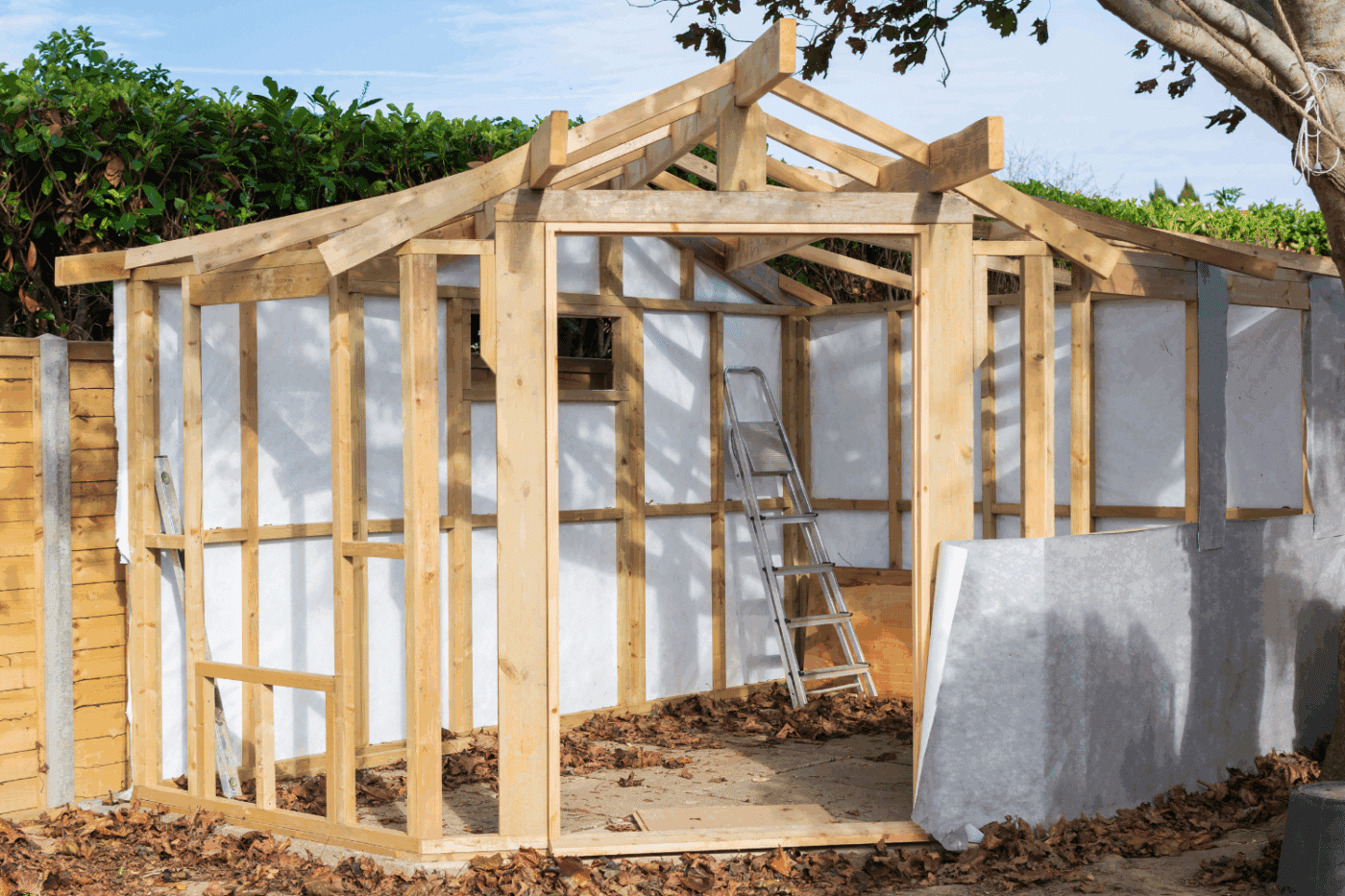 framed out shed in the corner of a yard. leaves are scattered inside and out the partially built shed