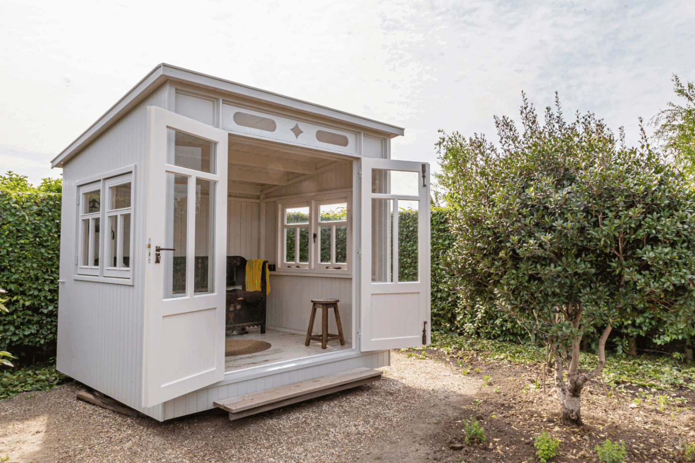 photo of a small white square studio shed with a center split door being fully open. shed sits in front of a hedge row