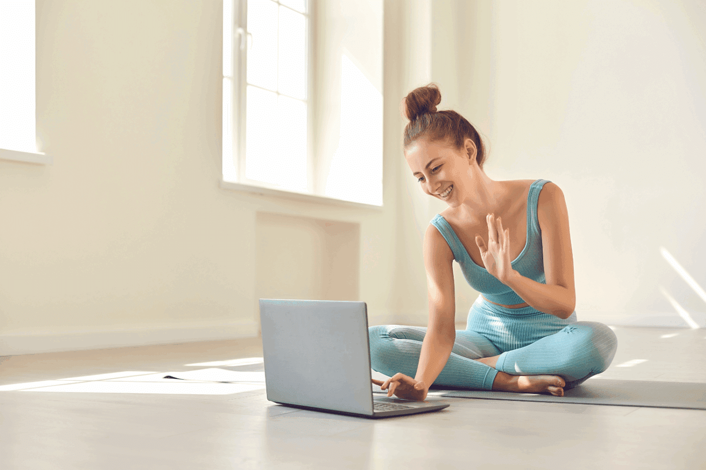 photo of a women in teal athletic wear sitting in an empty room on a yoga mat waving to an open laptop