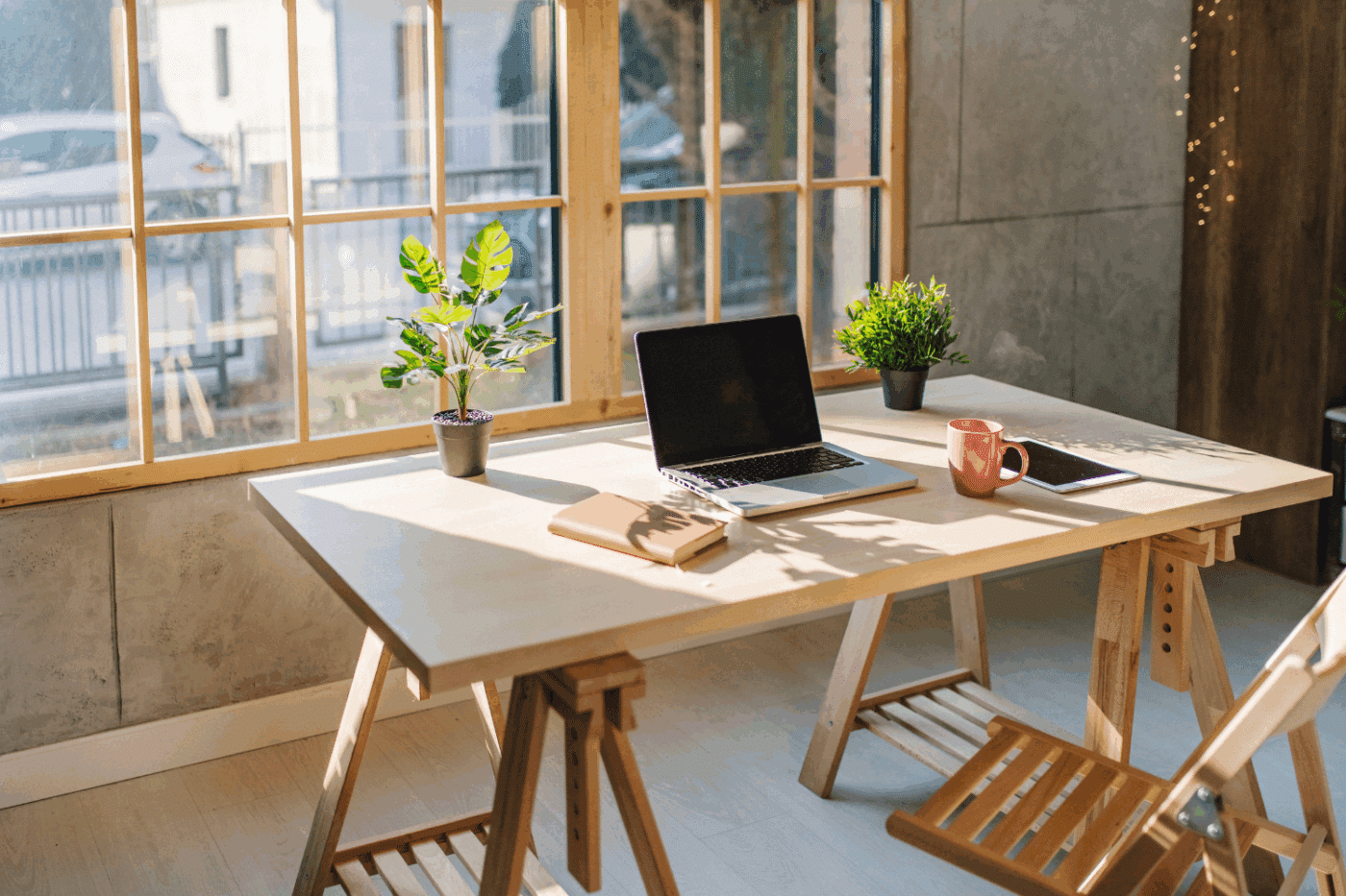 interior shot of a desk table in front of a window. with a laptop, mug, tablet notebook and two plants on it.