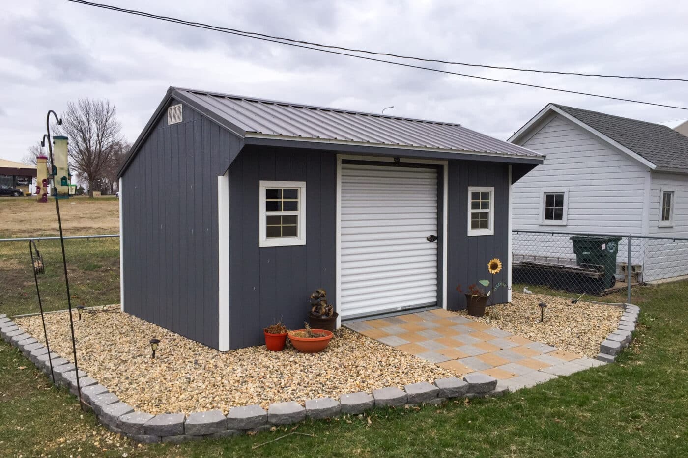 photo of a blue workshop shed with a sliding garage door in the middle. stones and small decorative lights with a sunflower focus the space around the shed.