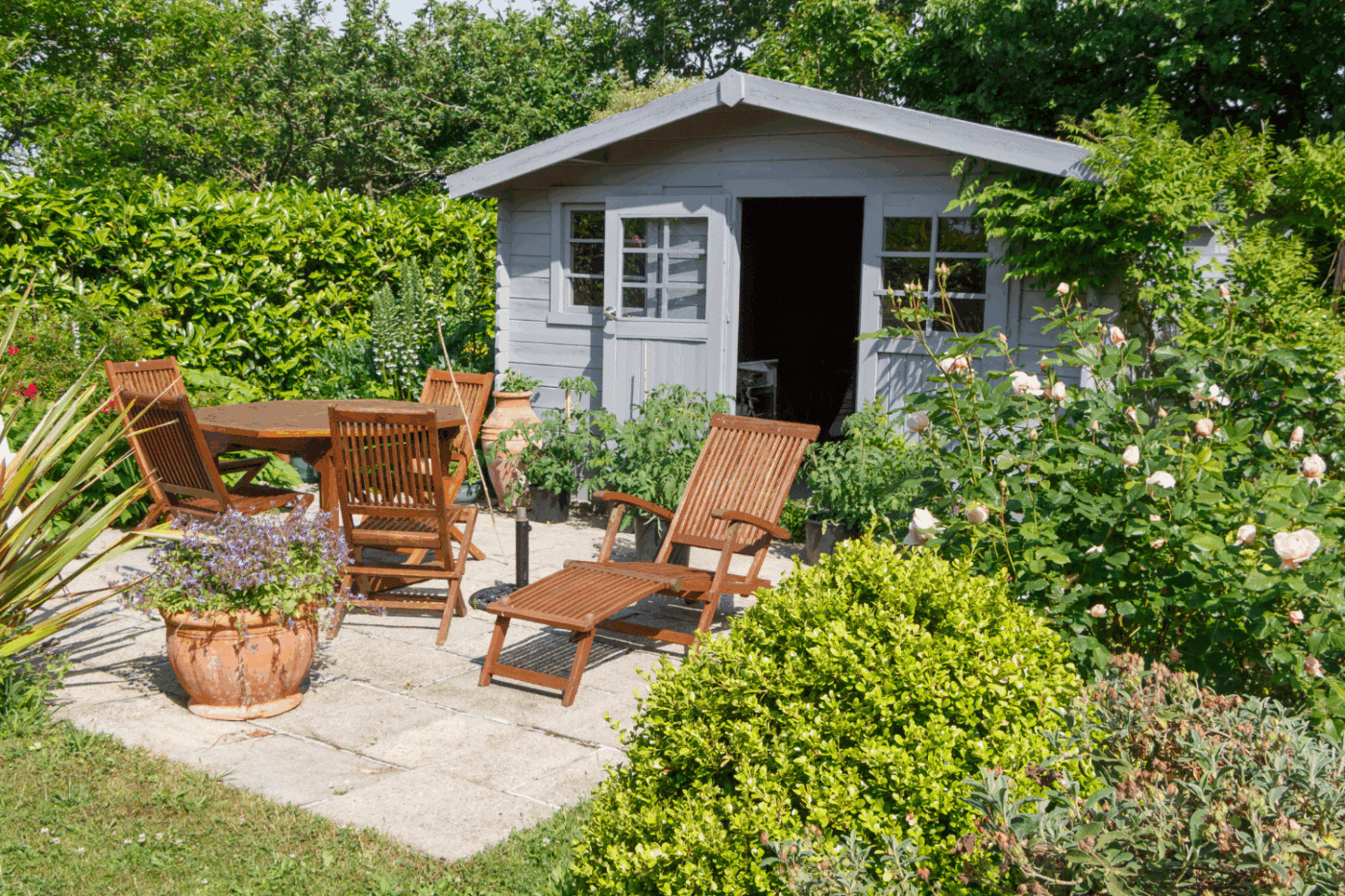 complete grey shed in front of a patio with lounge chairs and a table all surrounded by plants and flowers
